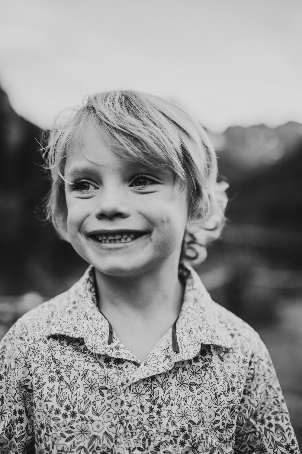 Black and white portrait of a young child with tousled hair wearing a floral print shirt and smiling brightly outdoors.
