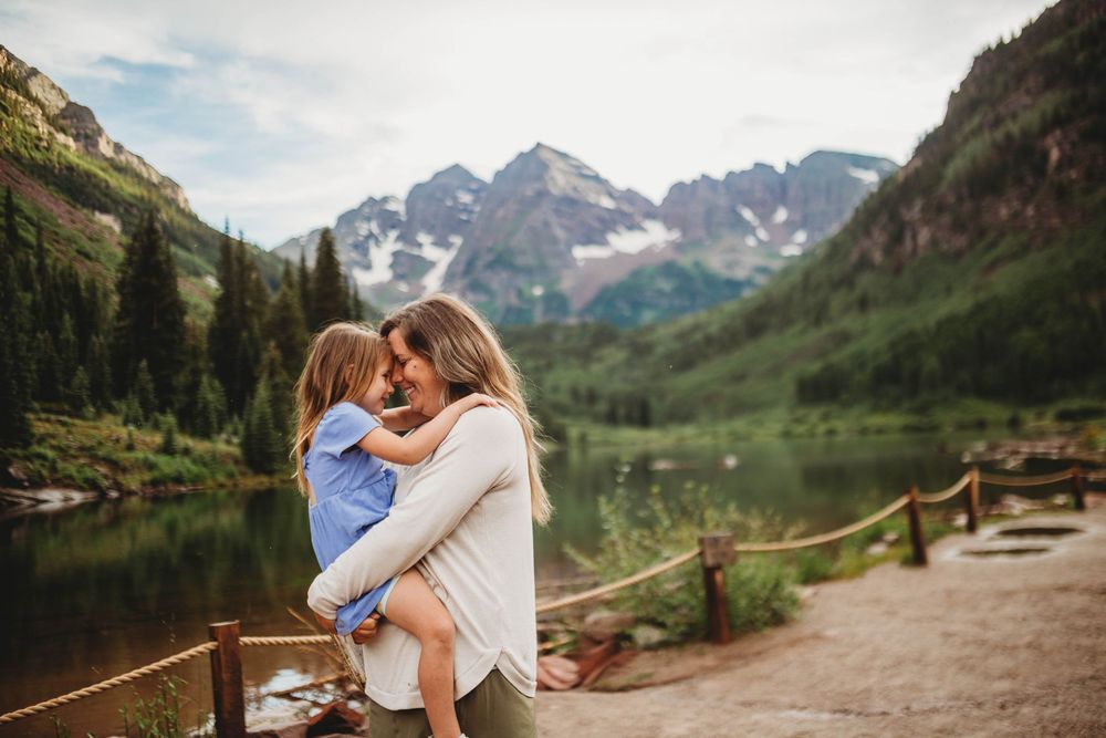 Two people share an embrace on a scenic mountain trail with snow-capped peaks and evergreen forests in the background.