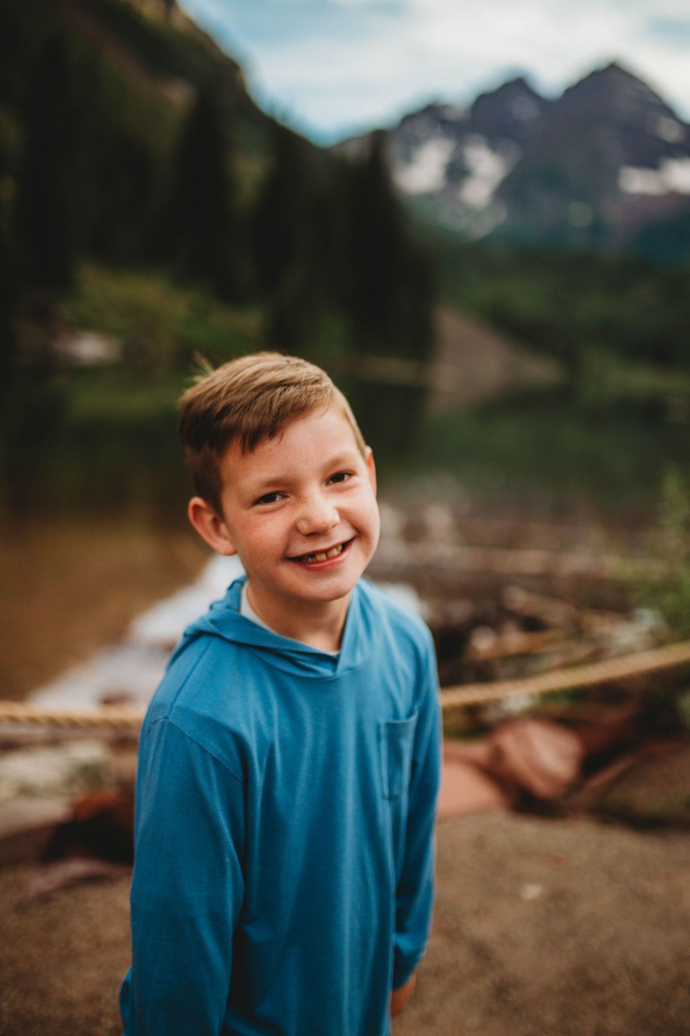 A child wearing a bright blue shirt and smiling outdoors with mountain views in the background.