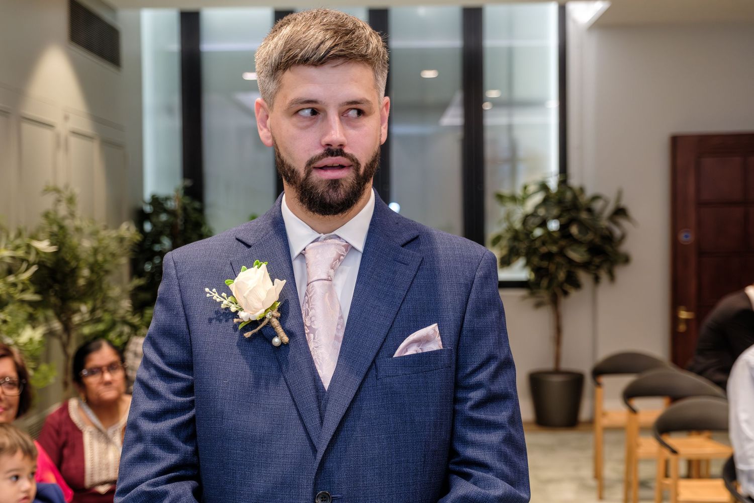 A groom in a navy suit with pink tie and white boutonniere stands at the altar during a wedding ceremony.
