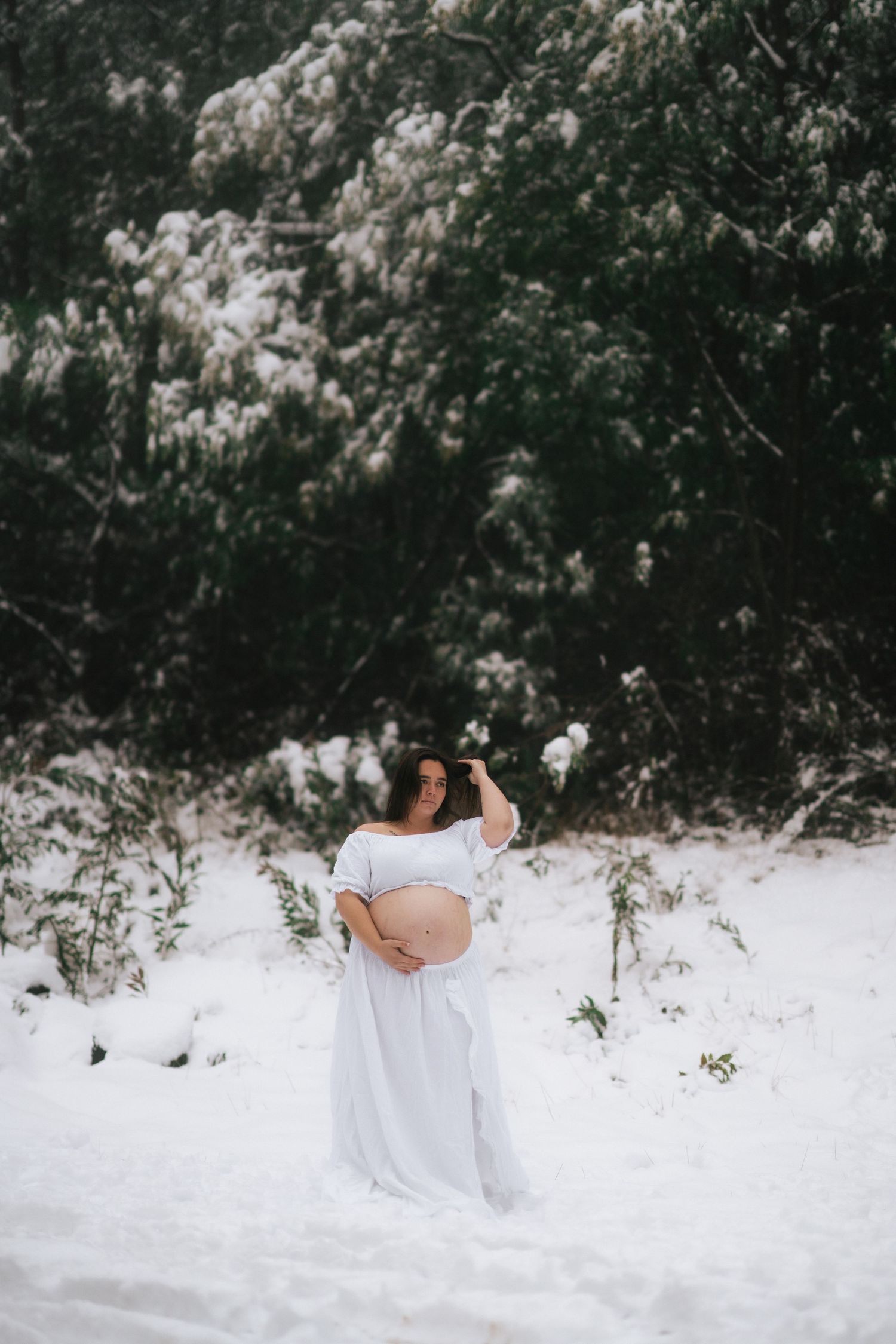 A maternity photo shoot in the snow features an expectant mother in a flowing white dress against a wintry backdrop.