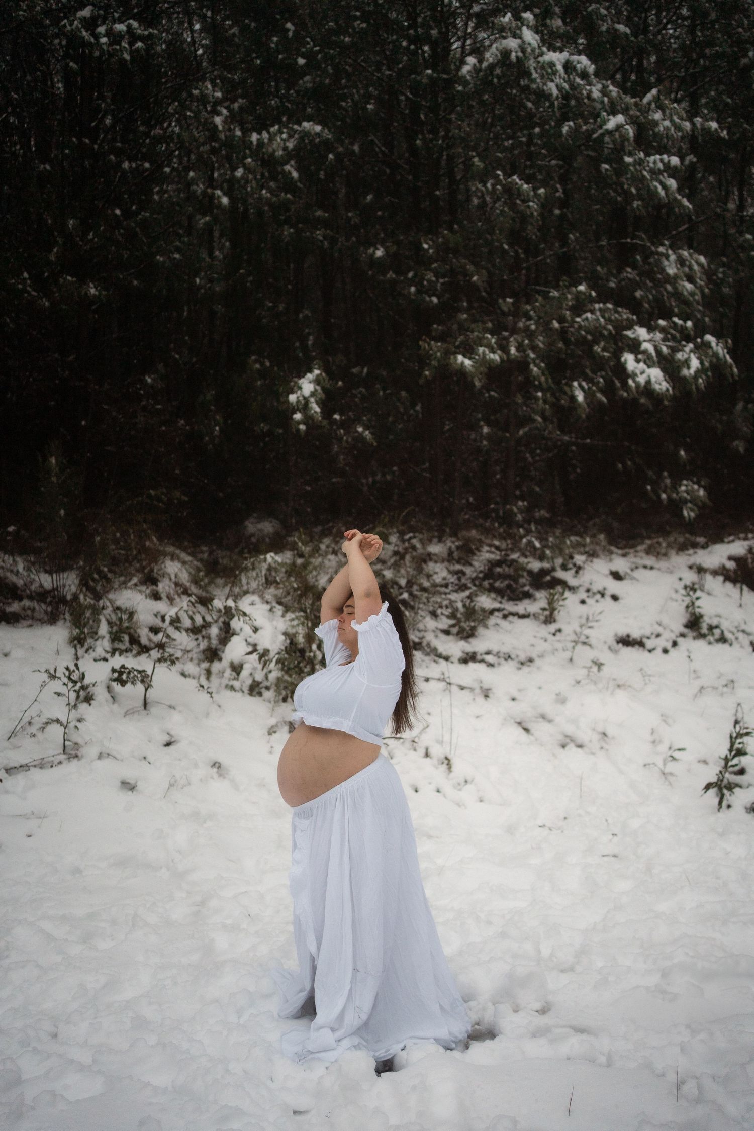 A bride in a white dress stands in snowy woods during a winter wedding photoshoot.