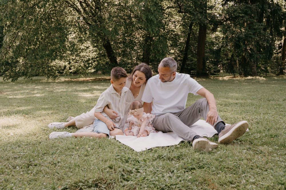 Familie entspannt gemeinsam auf einer weißen Decke in einer sonnigen Parklandschaft.