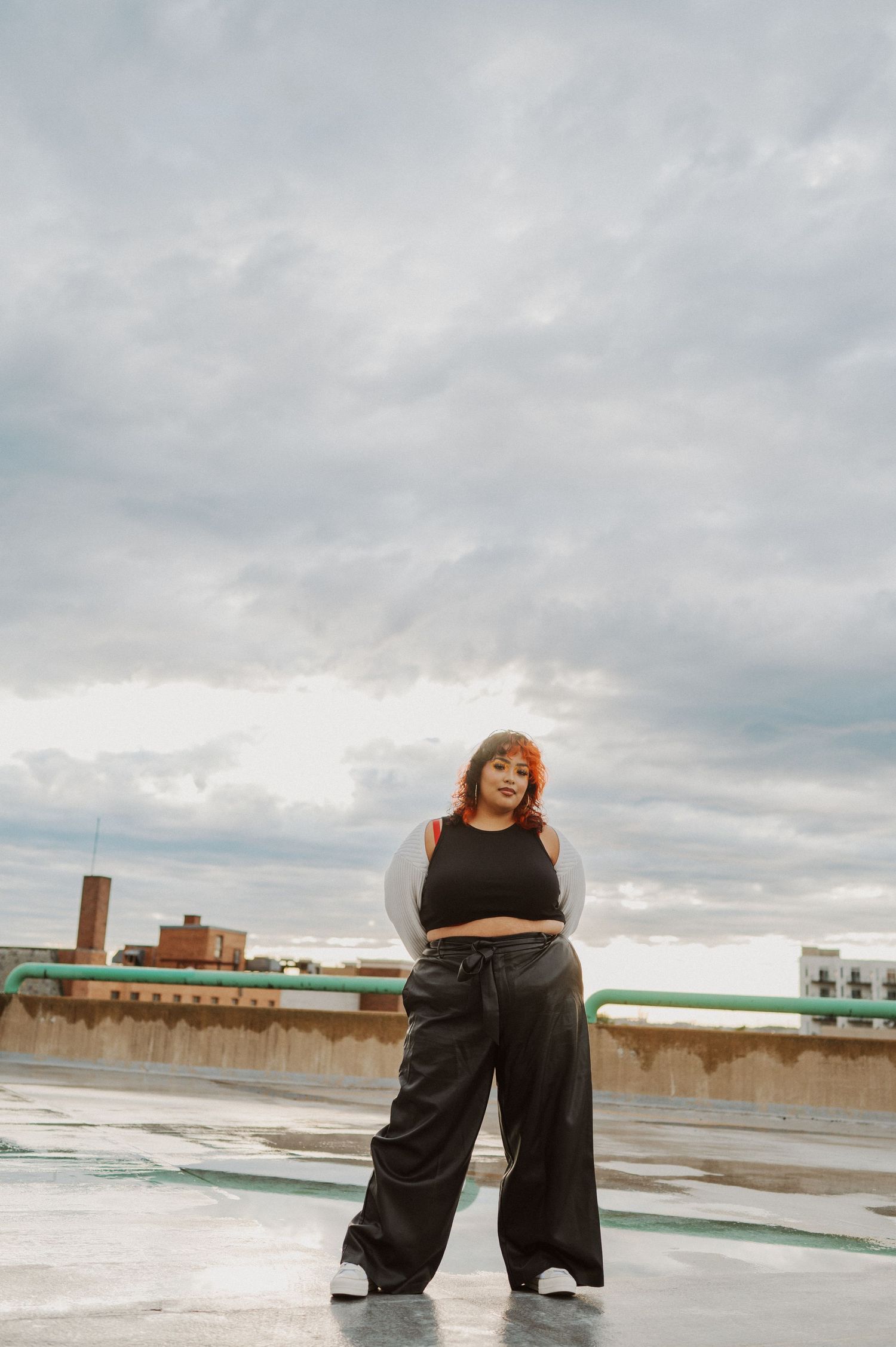 A person in a black crop top and wide-leg pants poses confidently on a rooftop under dramatic cloudy skies.