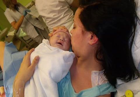 A newborn baby wrapped in a white blanket cries while being comforted in a medical setting with green walls.