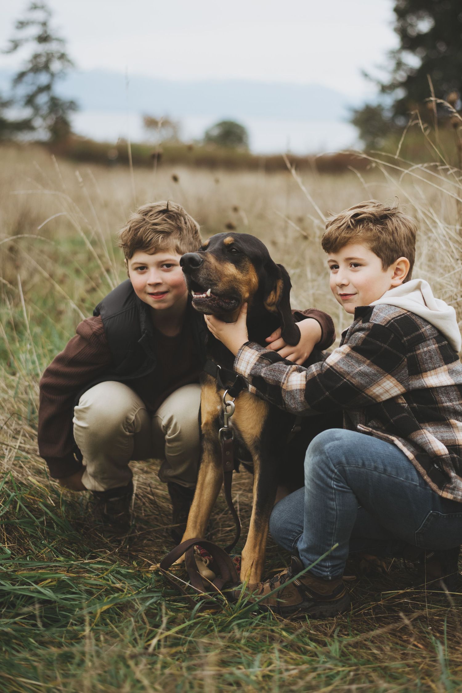 Two children kneel in a grassy field with their black and tan dog on an autumn day.