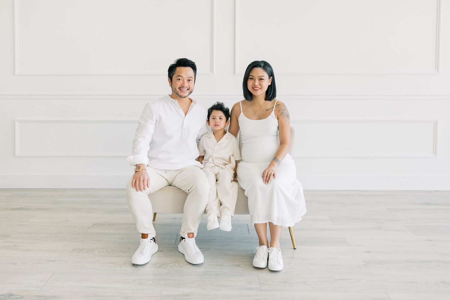 A family of three sits together in matching white outfits on a light gray floor.