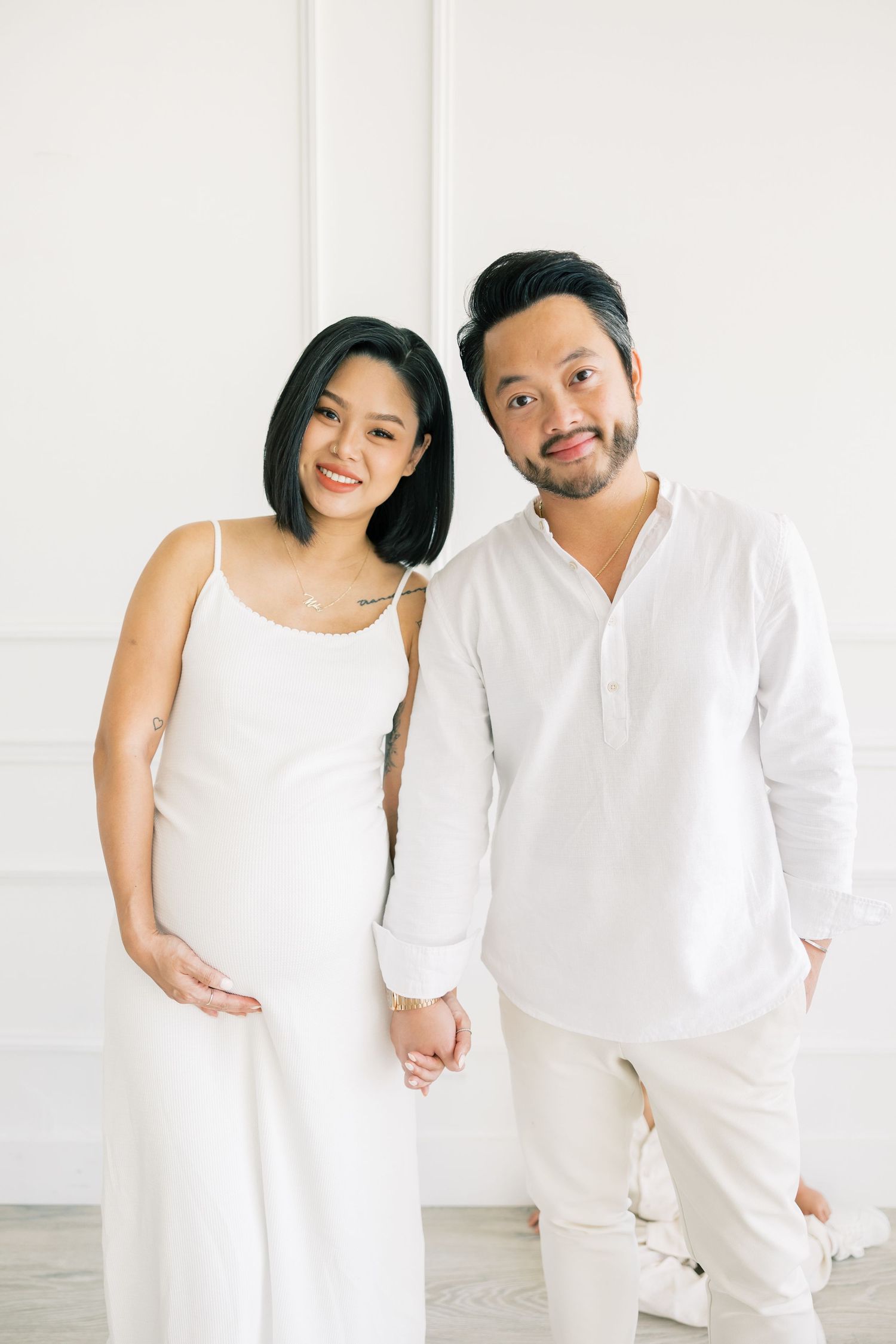 A couple in matching white outfits smiles warmly in a bright modern studio setting.