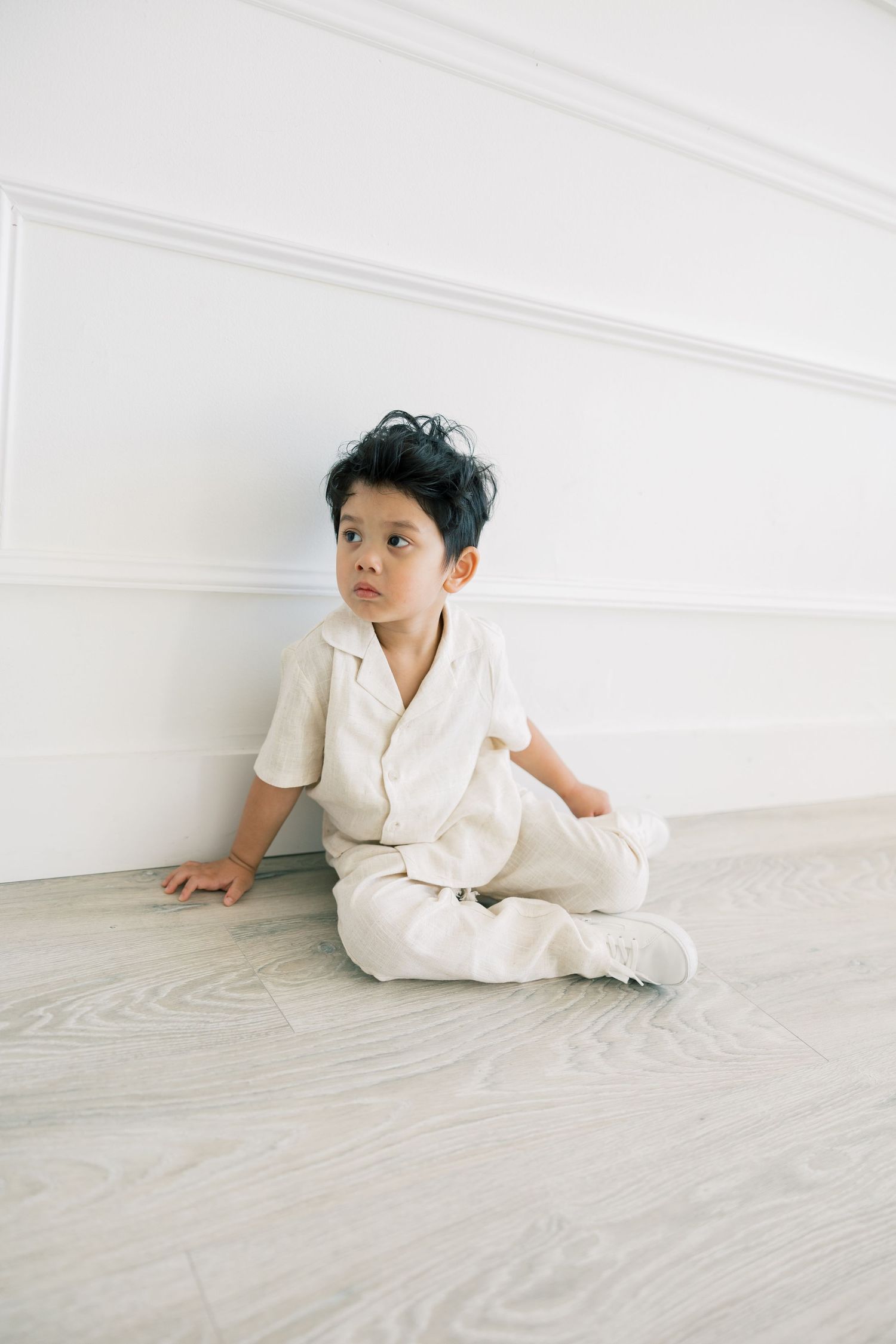 A young child in cream-colored clothing sits casually on a light wooden floor against a white wall.