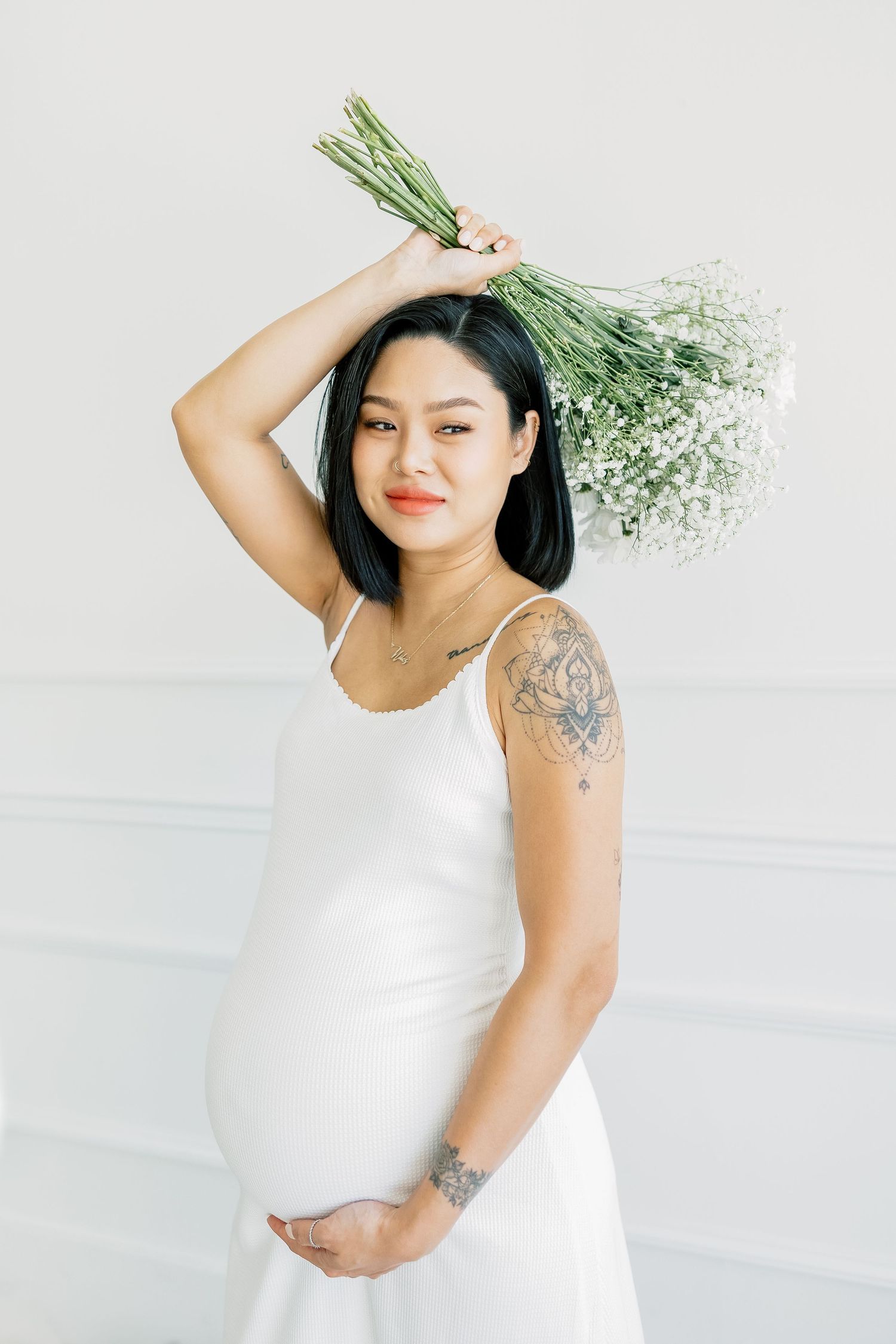 A pregnant woman in white dress poses playfully with baby's breath flowers held above her head.