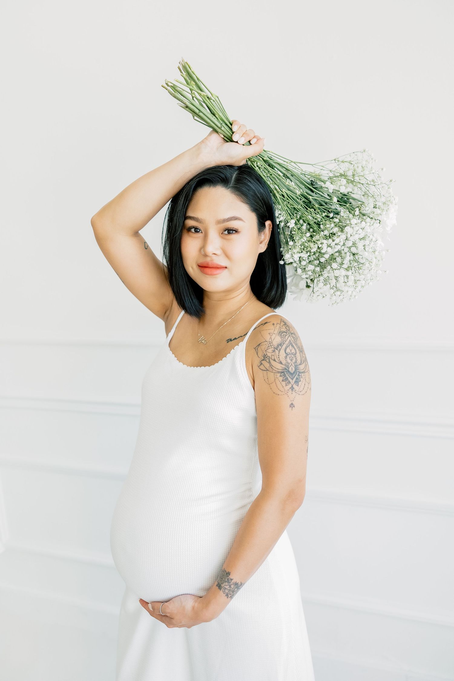 An expectant mother in a sleeveless white dress holds flowers above her head while cradling her belly.