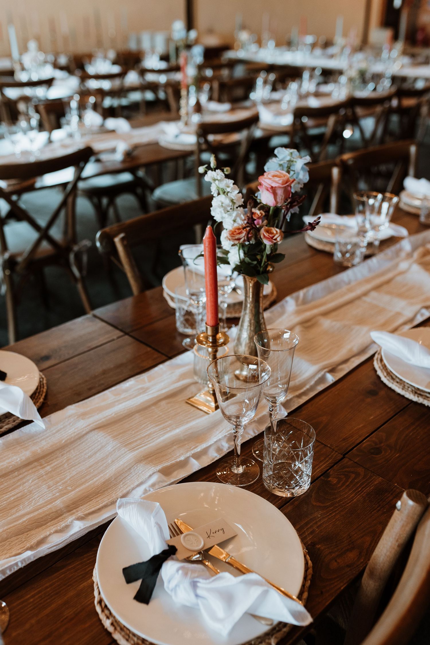 Rustic wedding reception table with white plates, coral candles, floral centerpieces and farmhouse chairs set for dinner.