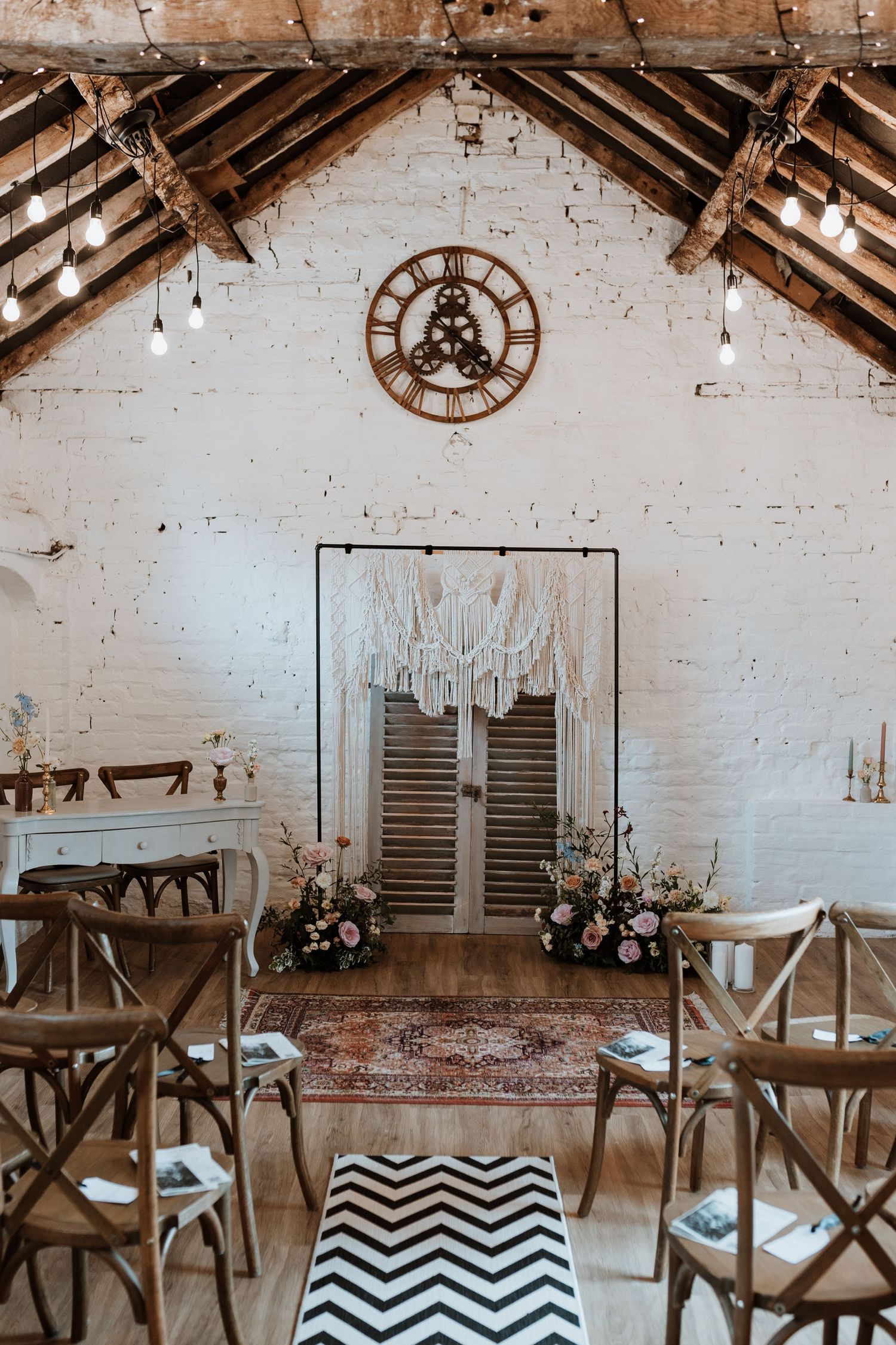 Rustic wedding ceremony setup with exposed wooden beams, white brick walls, and geometric rug leading to vintage doors.