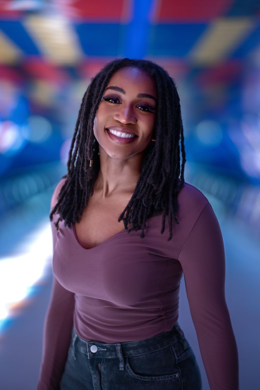 A person in a brown long-sleeve top and jeans smiles warmly against a blurred blue background.