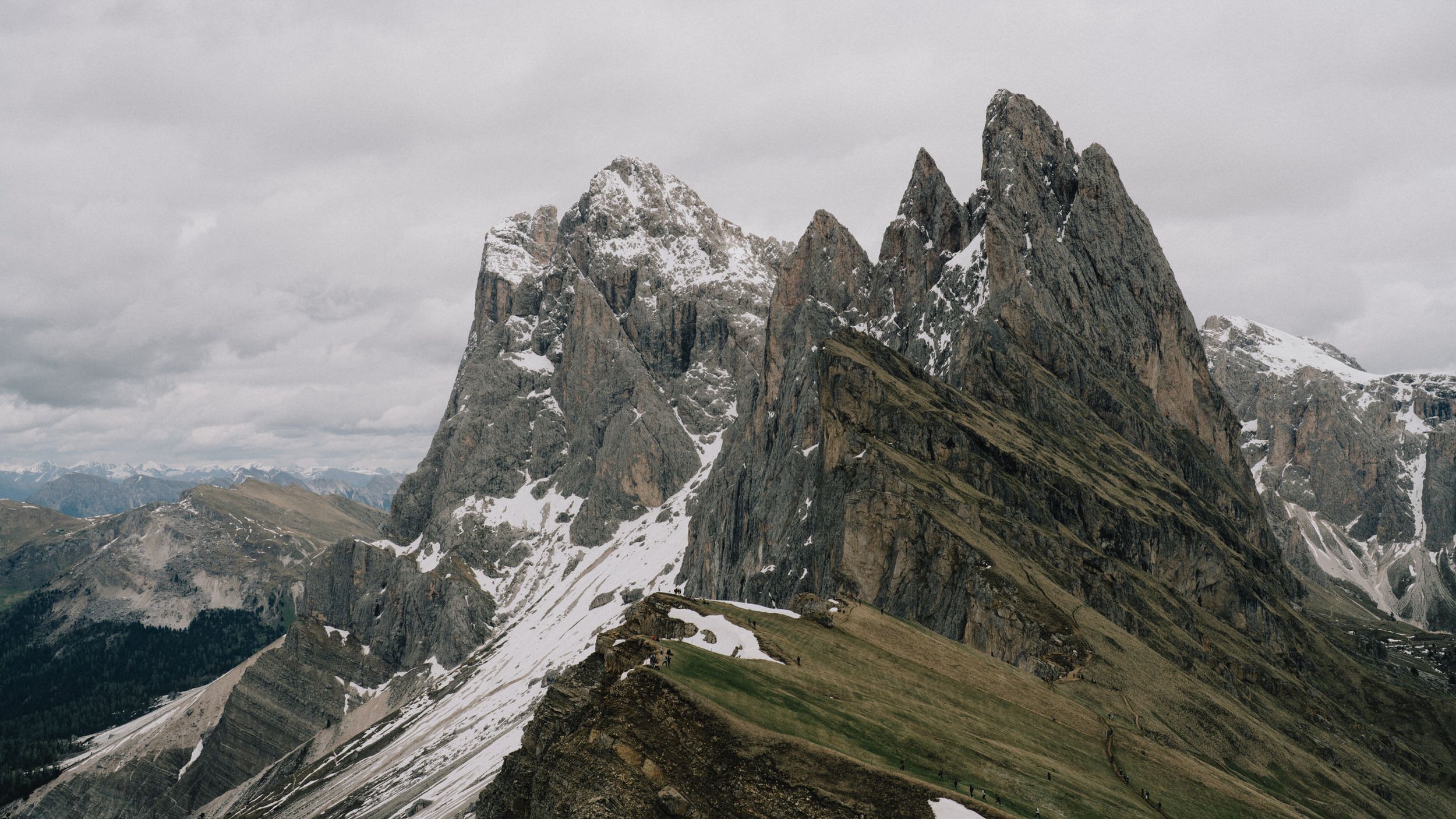 Dramatic panoramic view of snow-capped mountain peaks with jagged rock formations under cloudy skies.