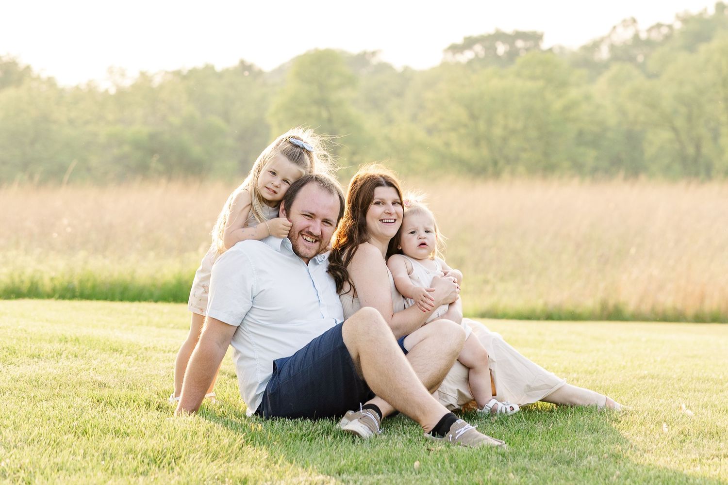 A family sits together on grass in a peaceful meadow during golden hour with tall grasses and trees in background.