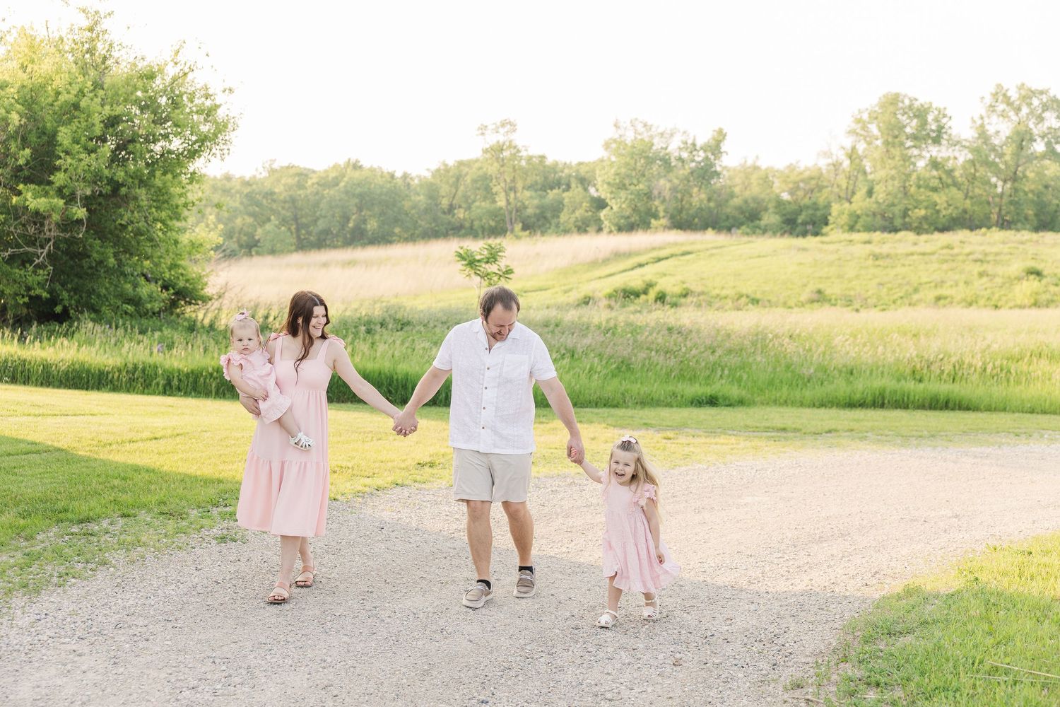 People holding hands while walking along a gravel path surrounded by green summer fields and trees.