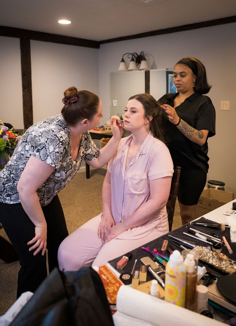 Makeup artists work on styling someone in a pink outfit while surrounded by beauty products and tools on a table.
