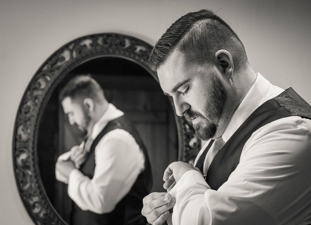 A bearded man in a white dress shirt and vest stands beside an ornate mirror, adjusting his tie in a black and white photo.