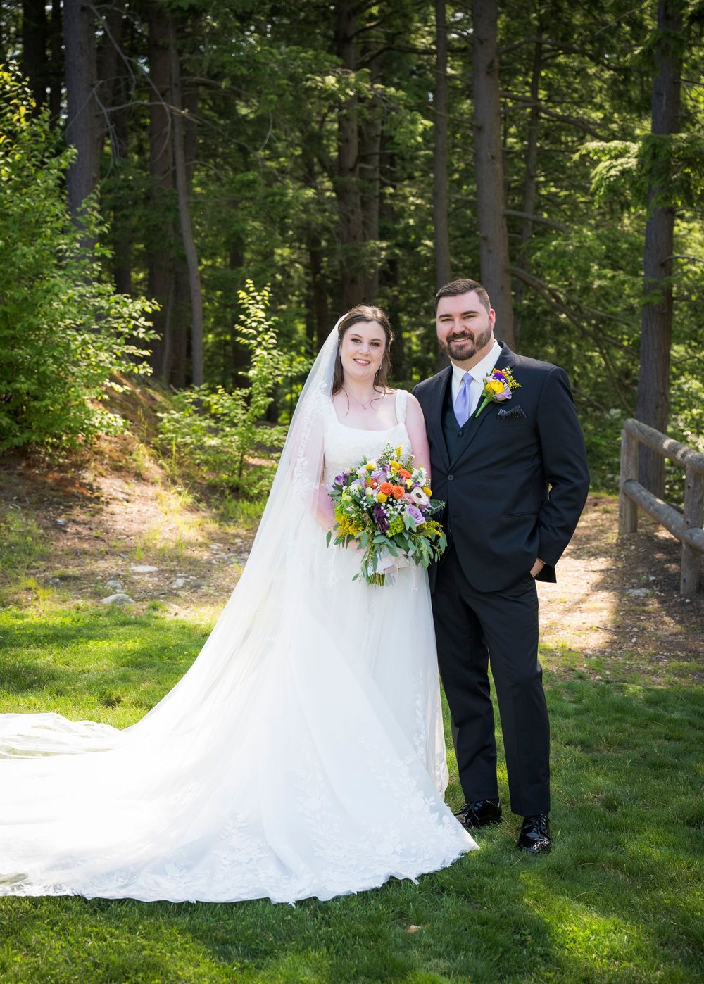 A couple poses together on a grassy hillside surrounded by tall evergreen trees.