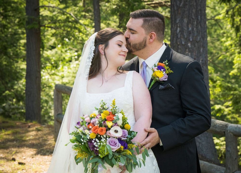 Romantic wedding couple embraces in a sunlit forest setting with colorful floral bouquet and formal attire.