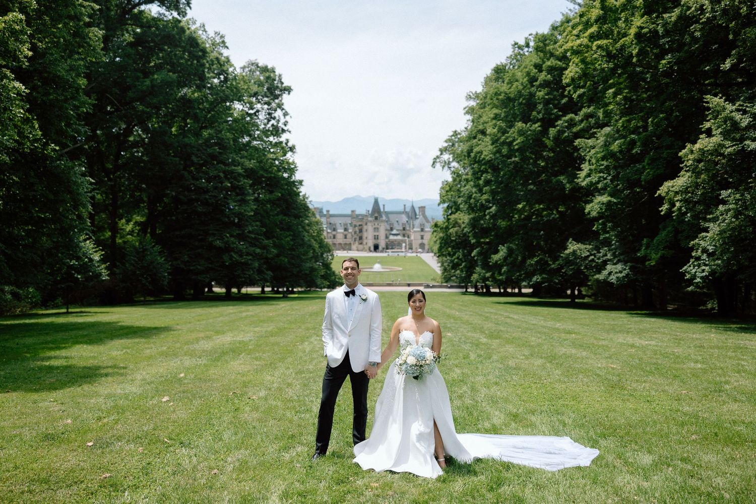 Elegant wedding couple stands on manicured lawn with grand mansion and trees in background on sunny day.