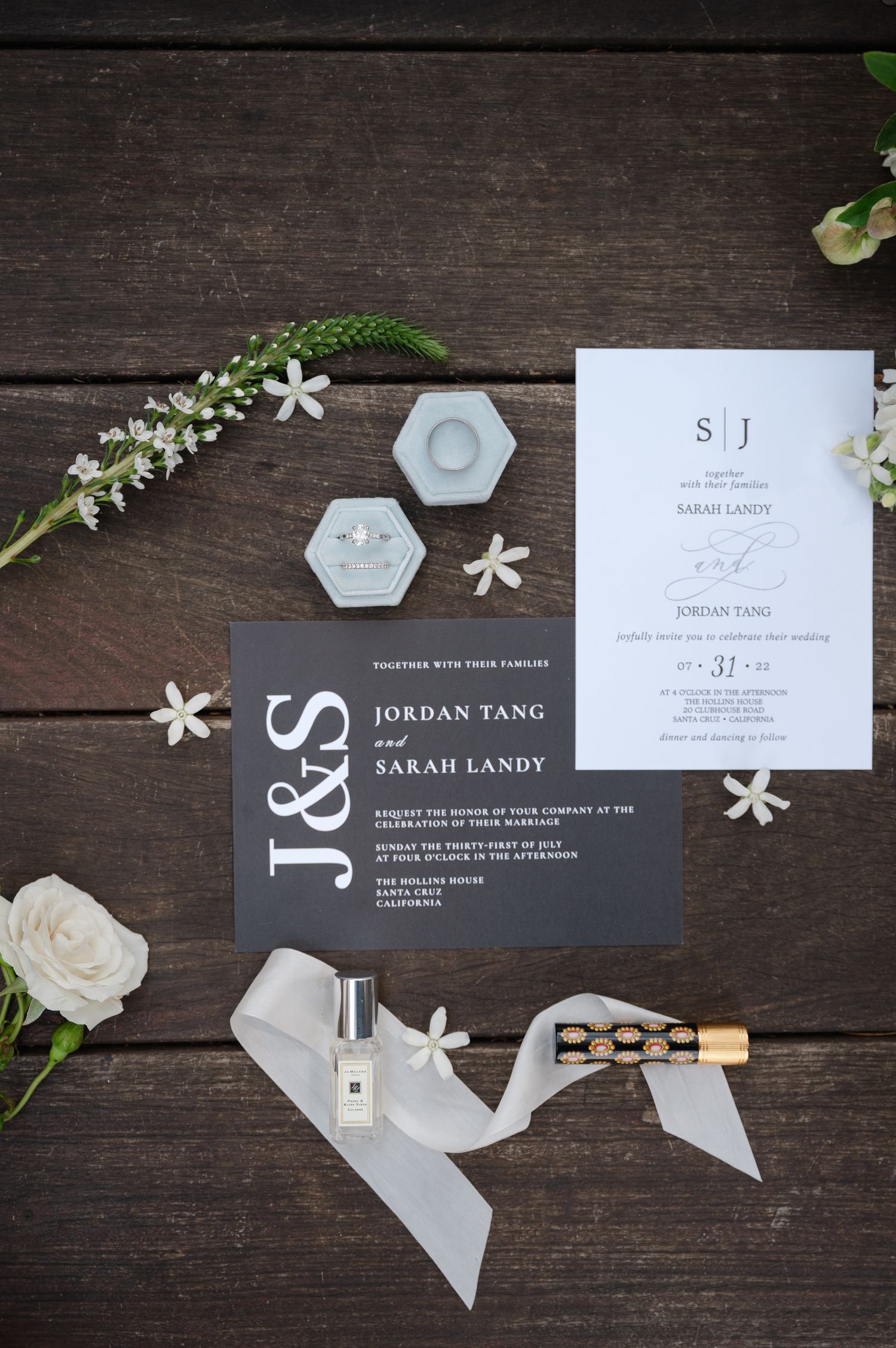 Elegant wedding invitation suite with gray and white cards, hexagonal ring dishes, and delicate white flowers on dark wood.
