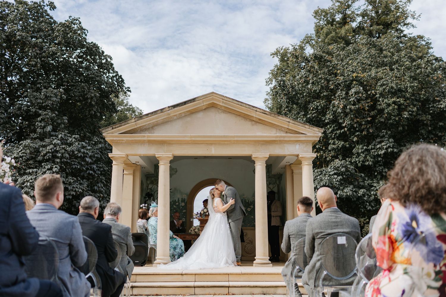 Outdoor wedding ceremony taking place at an elegant yellow gazebo with white columns surrounded by lush trees.