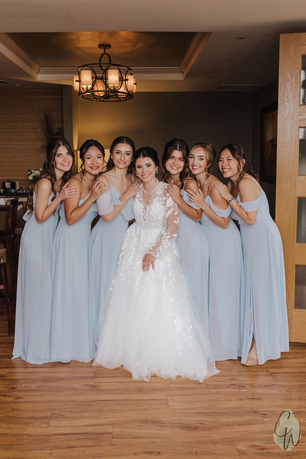 Bride in white lace gown poses with bridesmaids wearing light blue dresses in an elegant indoor setting.