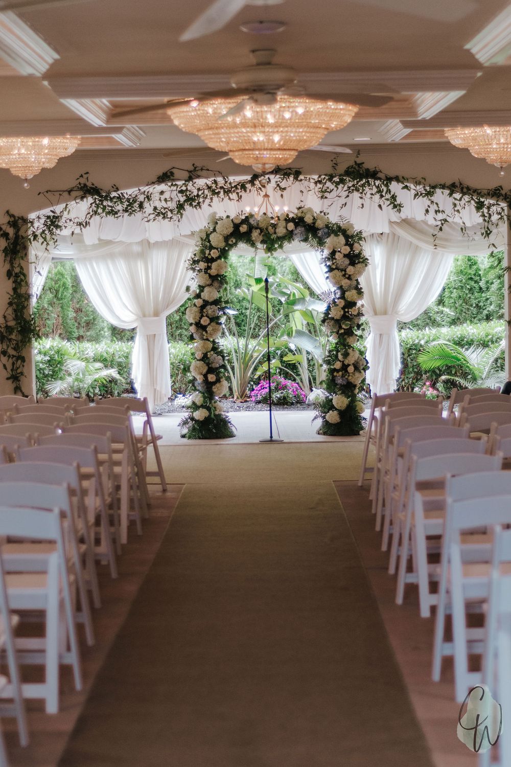 Indoor wedding venue decorated with white drapes, chandeliers, and floral arches along a carpeted aisle lined with white chairs.
