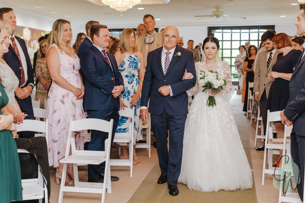 Two people walking down the aisle during an indoor wedding ceremony as guests look on from white chairs.