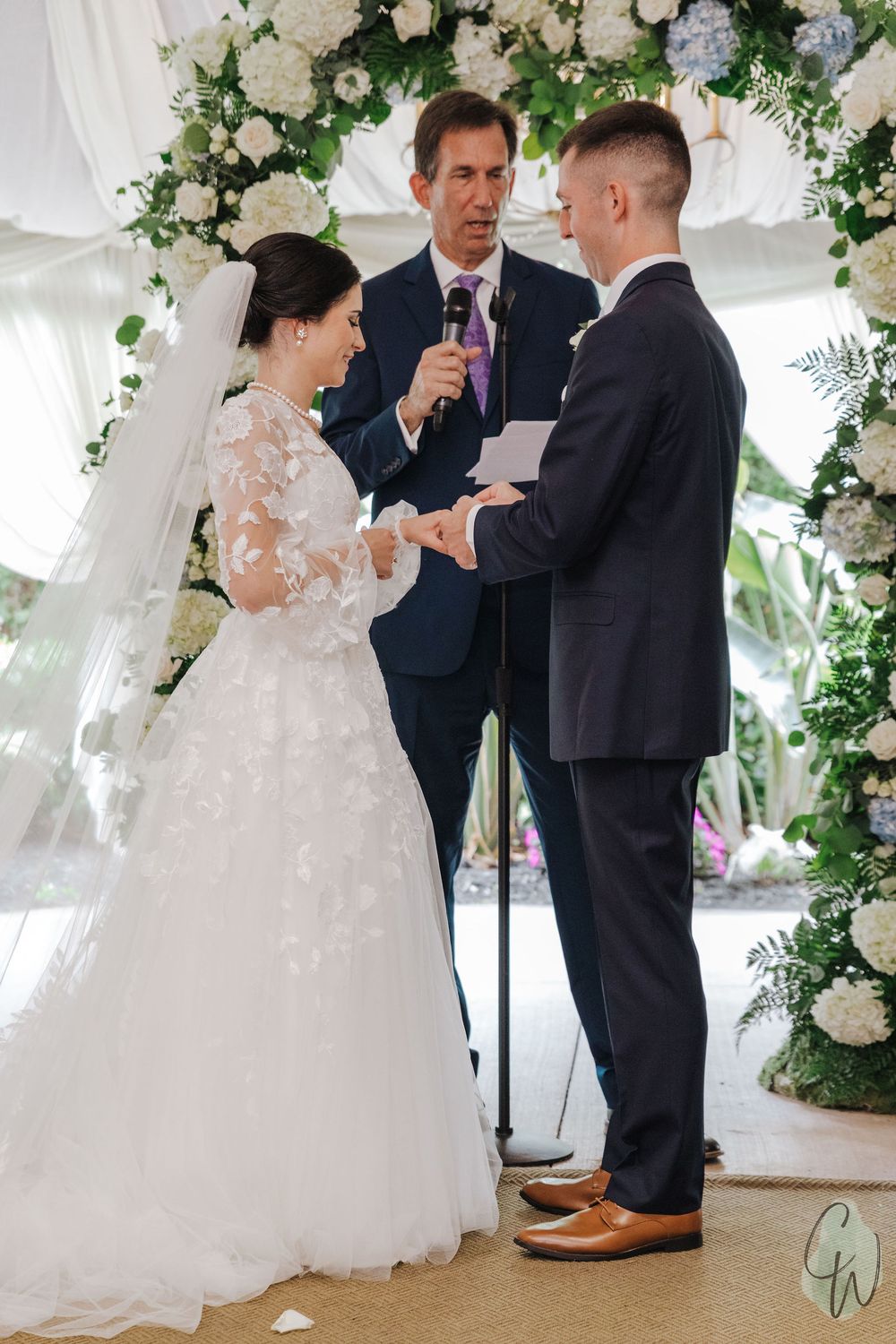 A wedding ceremony takes place under a white tent decorated with elegant floral arrangements and flowing white fabric.