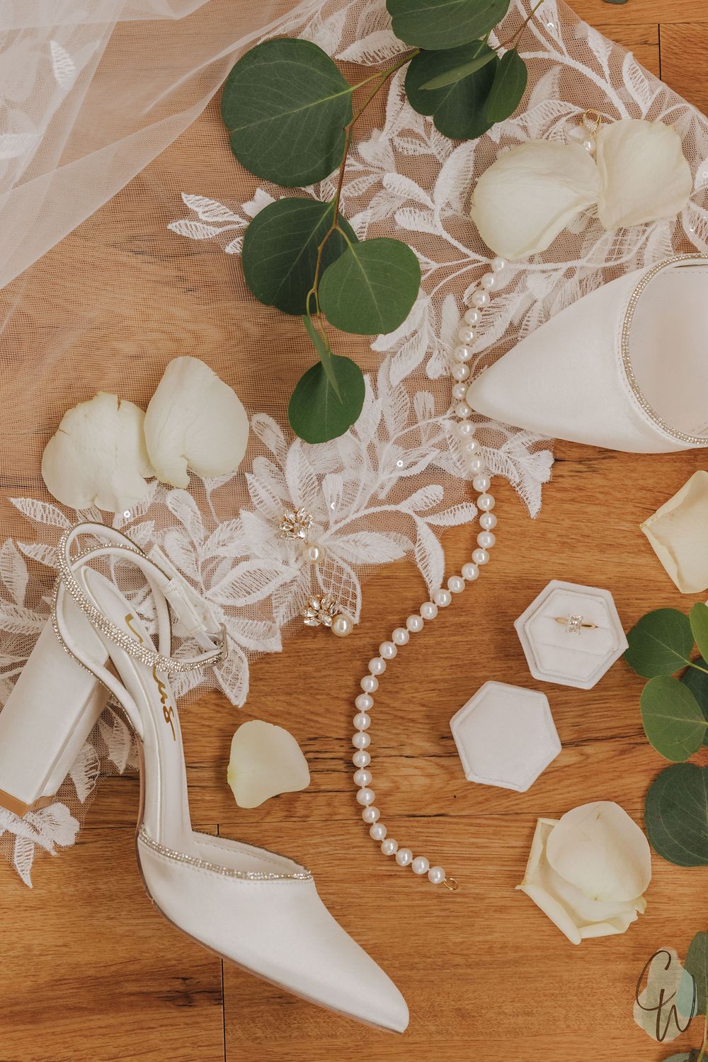 Wedding accessories including white shoes, pearls, floral elements and lace details arranged on wooden surface.