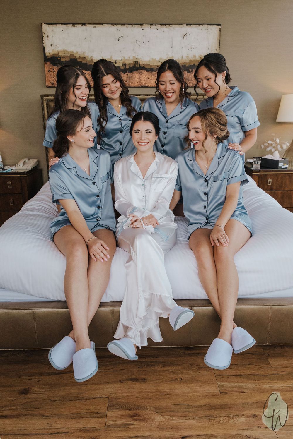A group of bridesmaids in matching blue pajama sets sitting with a bride in white on a bed, all wearing slippers.