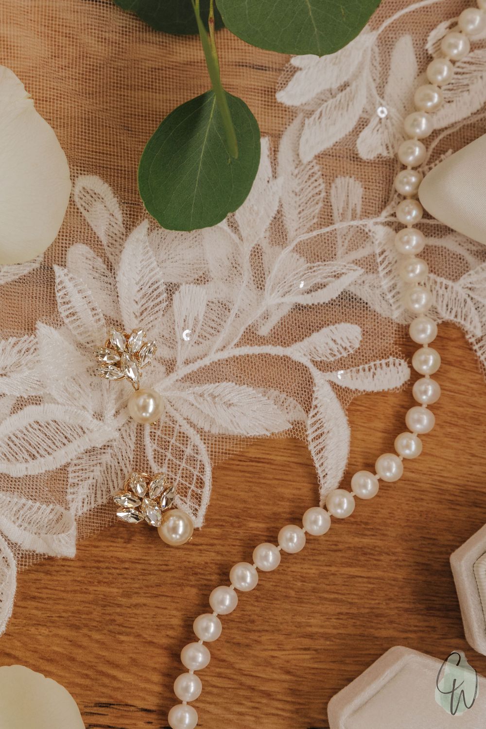 Wedding accessories including delicate white lace, pearl necklace, and crystal earrings arranged on wooden surface with green leaf.