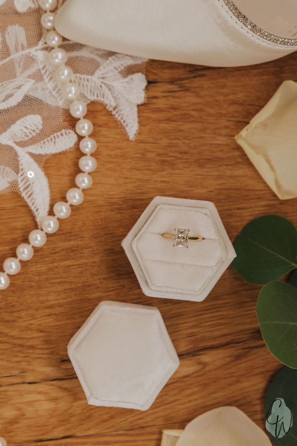 Elegant hexagonal ring box with pearl strands and lace details on wooden surface with green eucalyptus leaves.