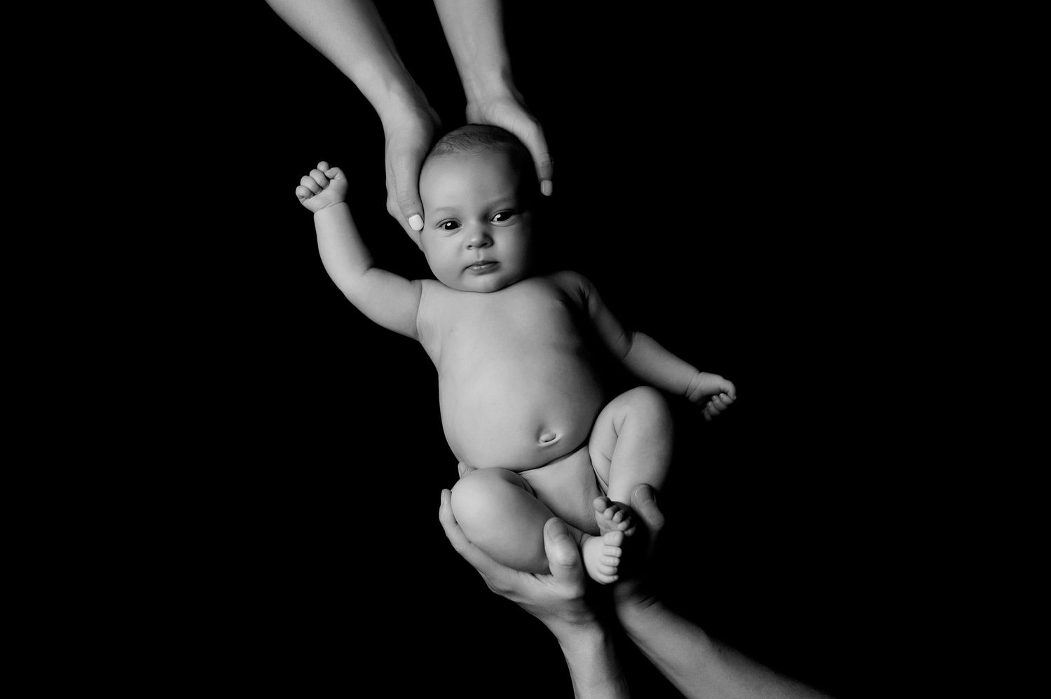 Black and white portrait of a baby wearing a diaper being supported by adult hands against dark background.