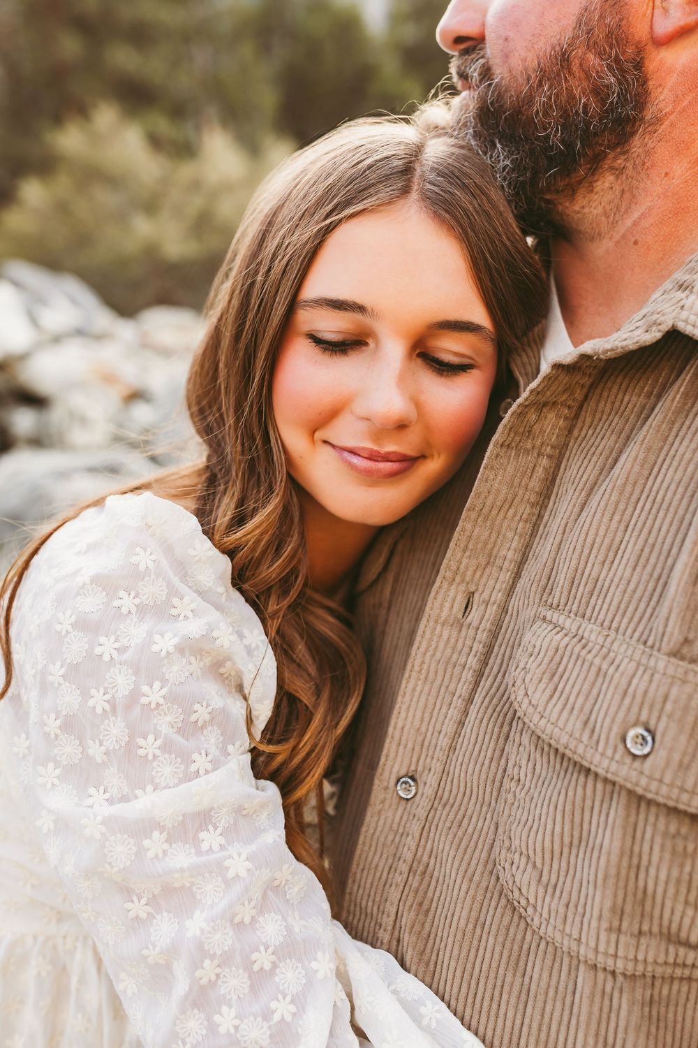 A romantic outdoor portrait showing a couple embracing, with her wearing a white lace dress and him in a corduroy shirt.