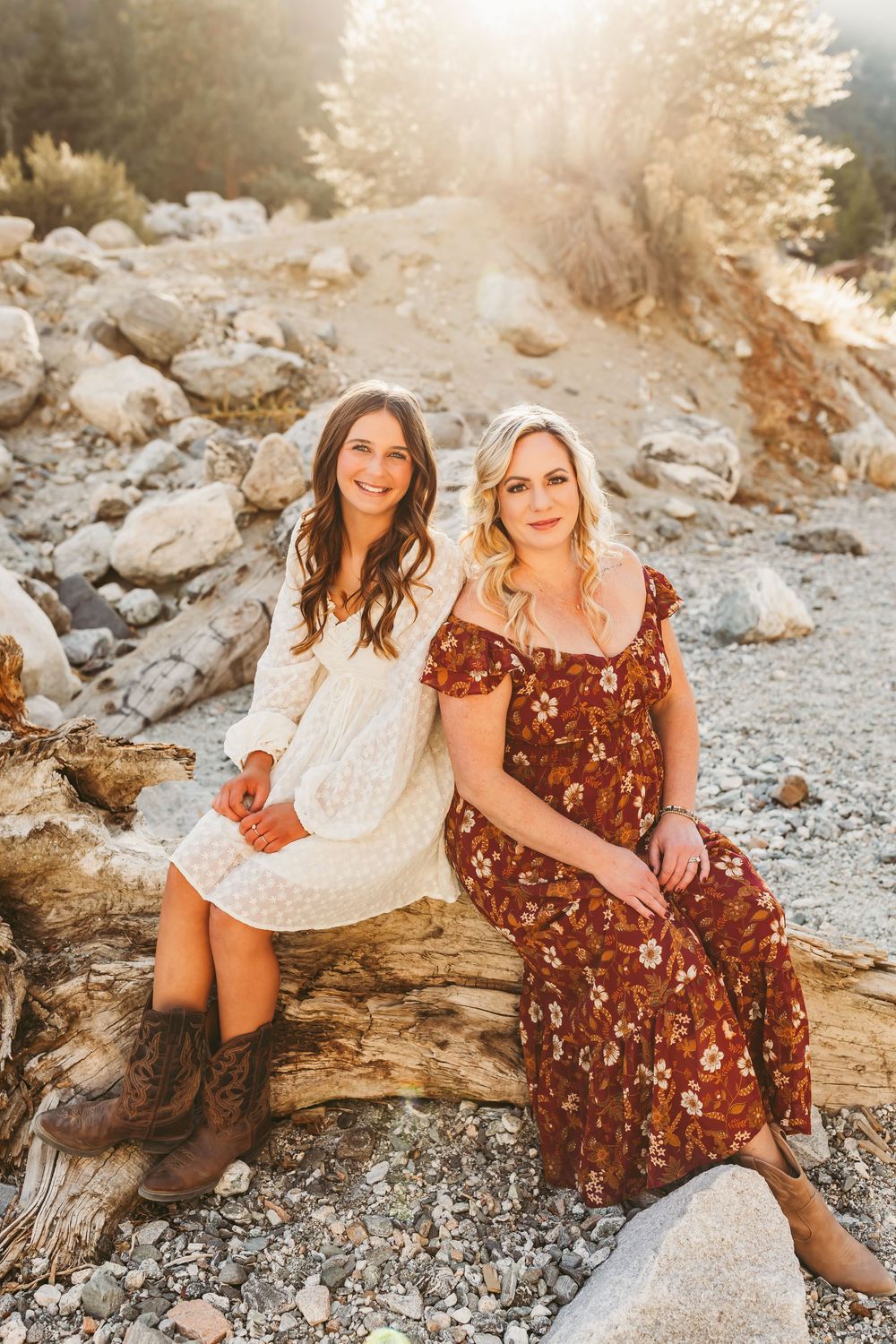 Two friends in floral dresses pose together on rocky terrain during golden hour sunset in nature.