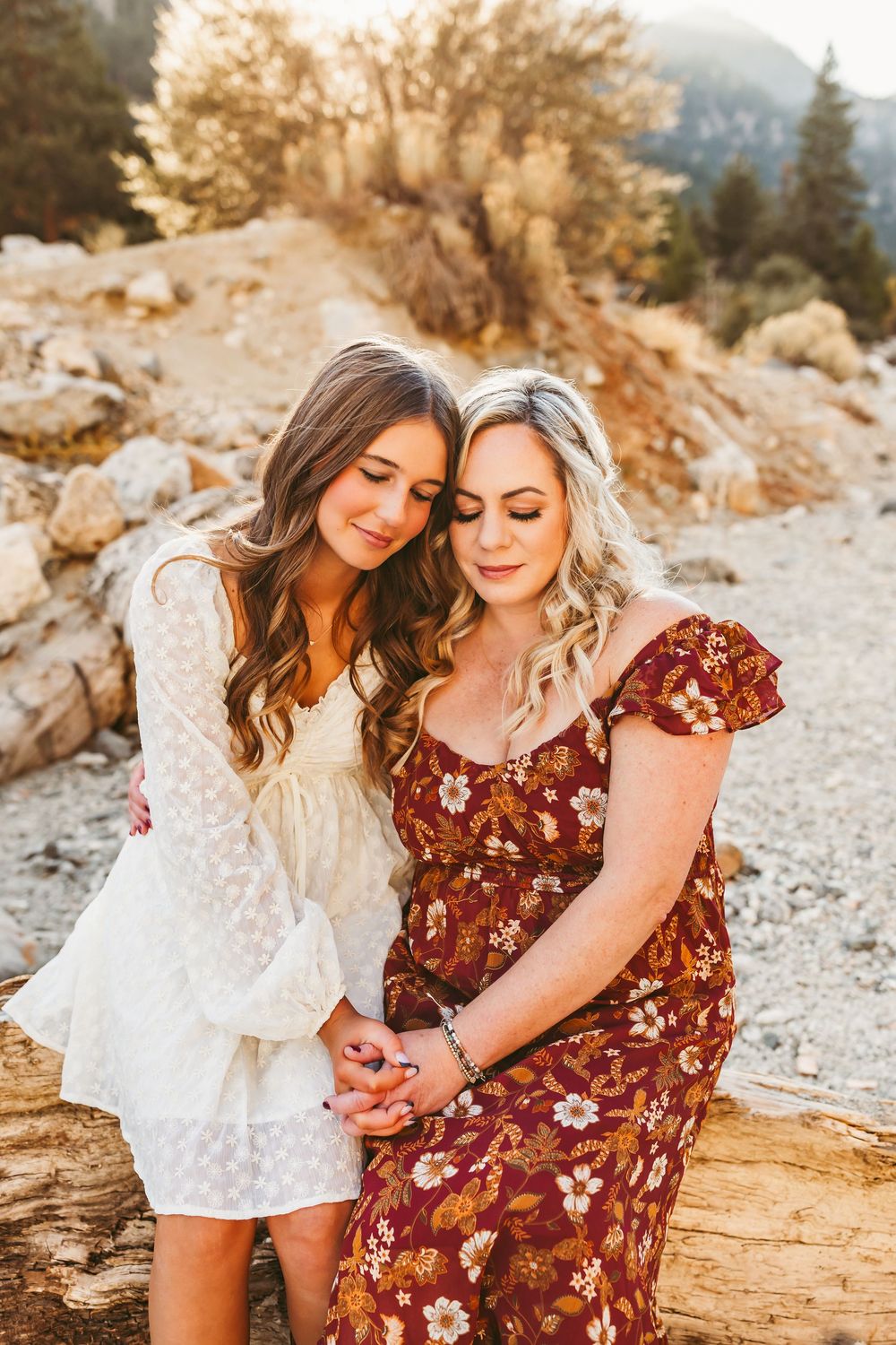 Two women in white and burgundy dresses sit close together on a fallen log in a sun-drenched mountain setting.