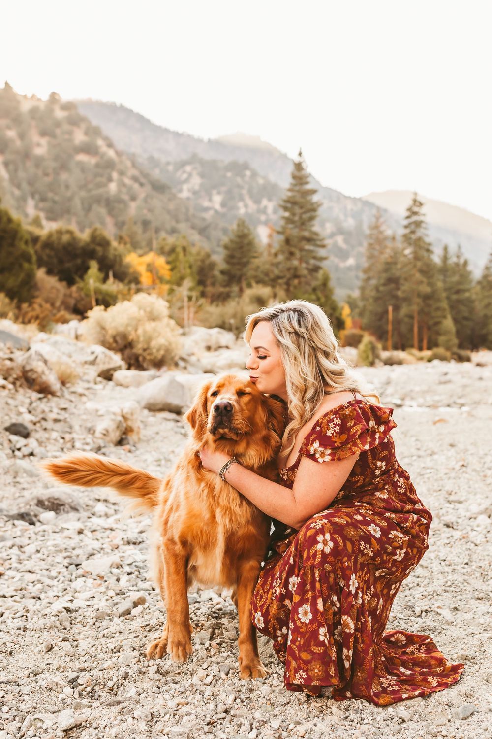 Woman in floral burgundy dress affectionately playing with golden retriever dog against mountain backdrop sunset.