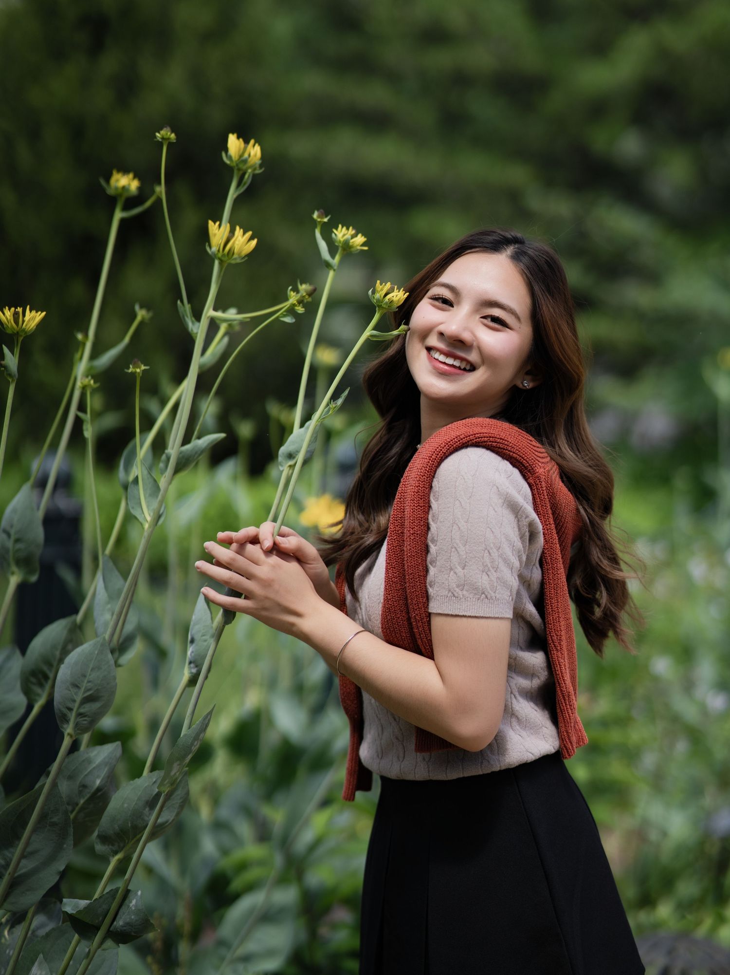 Portrait in a garden shows a happy expression while reaching out to touch yellow wildflowers against a blurred green background.