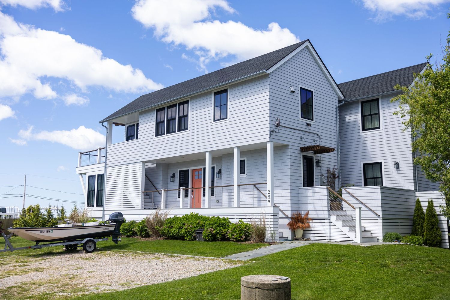 Modern white two-story coastal home with wraparound porch, red front door, and landscaped yard under blue skies.