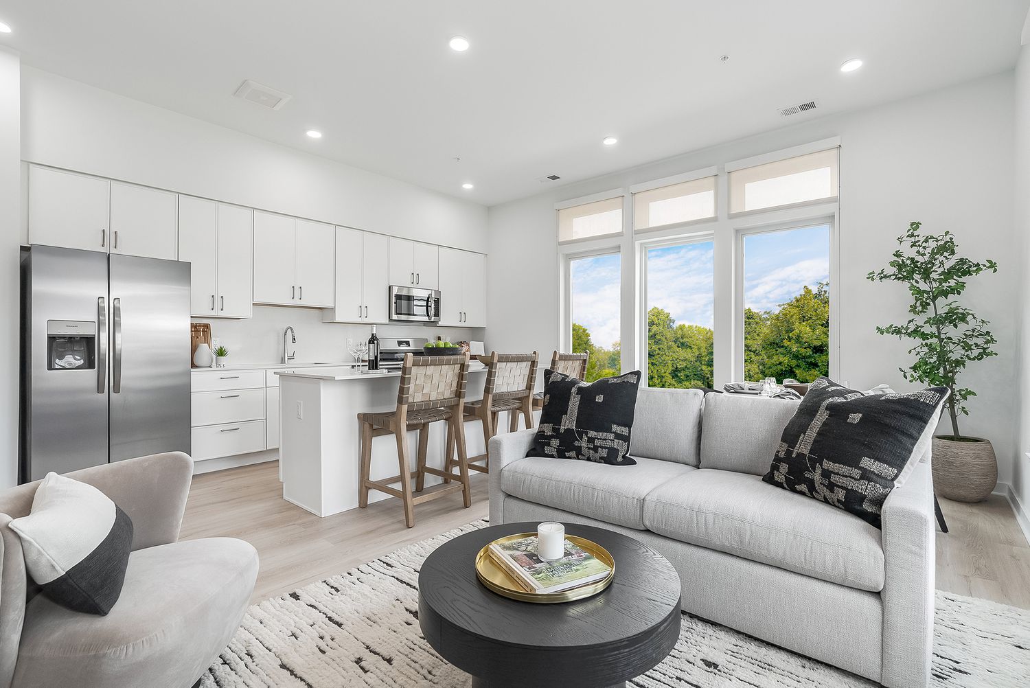 Modern open concept living room and kitchen with white cabinets, gray sectional sofa, and tall windows overlooking greenery.