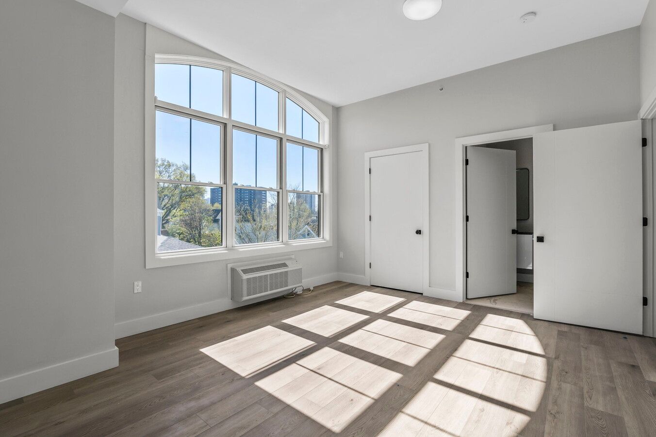 Bright modern bedroom with large arched window, sunlight casting geometric shadows on grey hardwood floors.