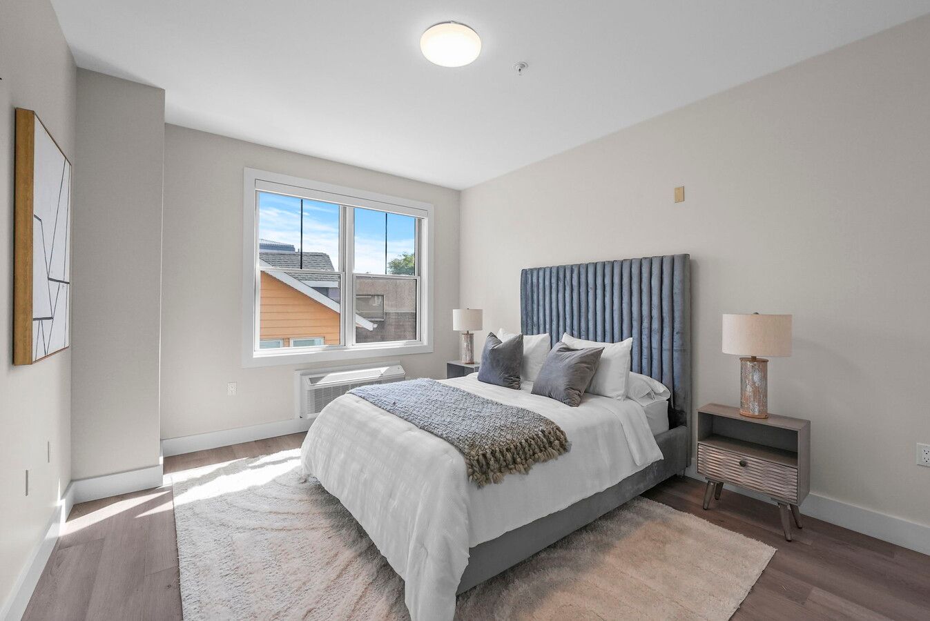 Modern bedroom with blue velvet headboard, white bedding, grey walls, and natural light from window.