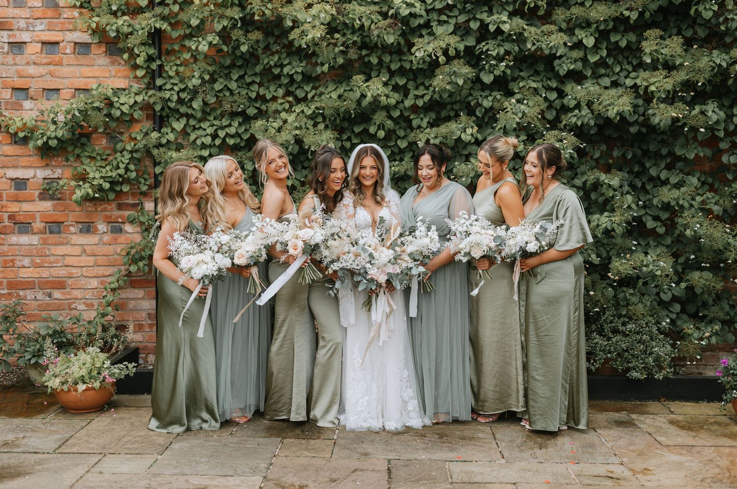 Bridal party in sage green dresses poses outdoors along brick wall for wedding photos.