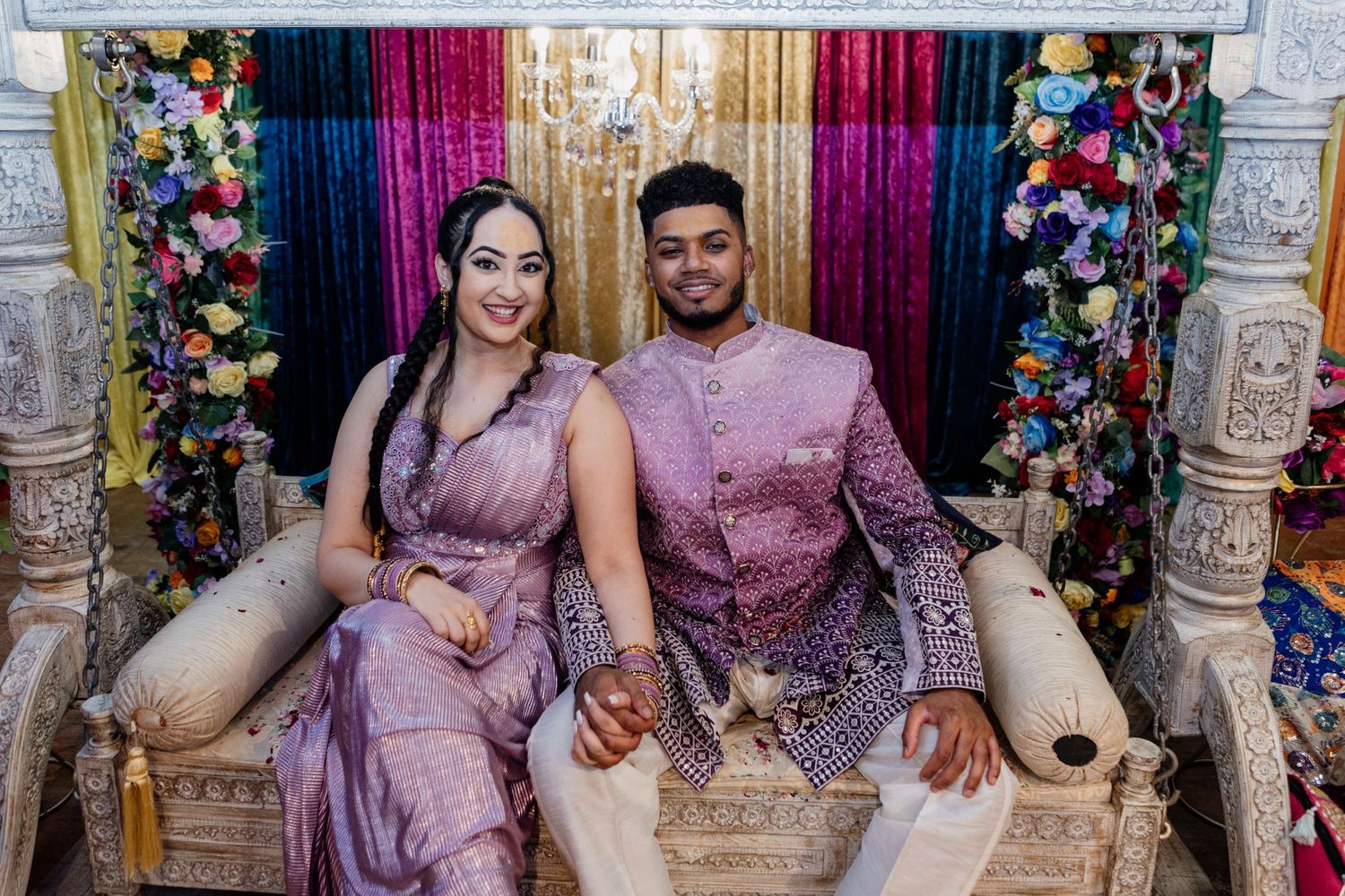 Couple in coordinating lavender and purple traditional outfits sit together at wedding celebration with floral backdrop.