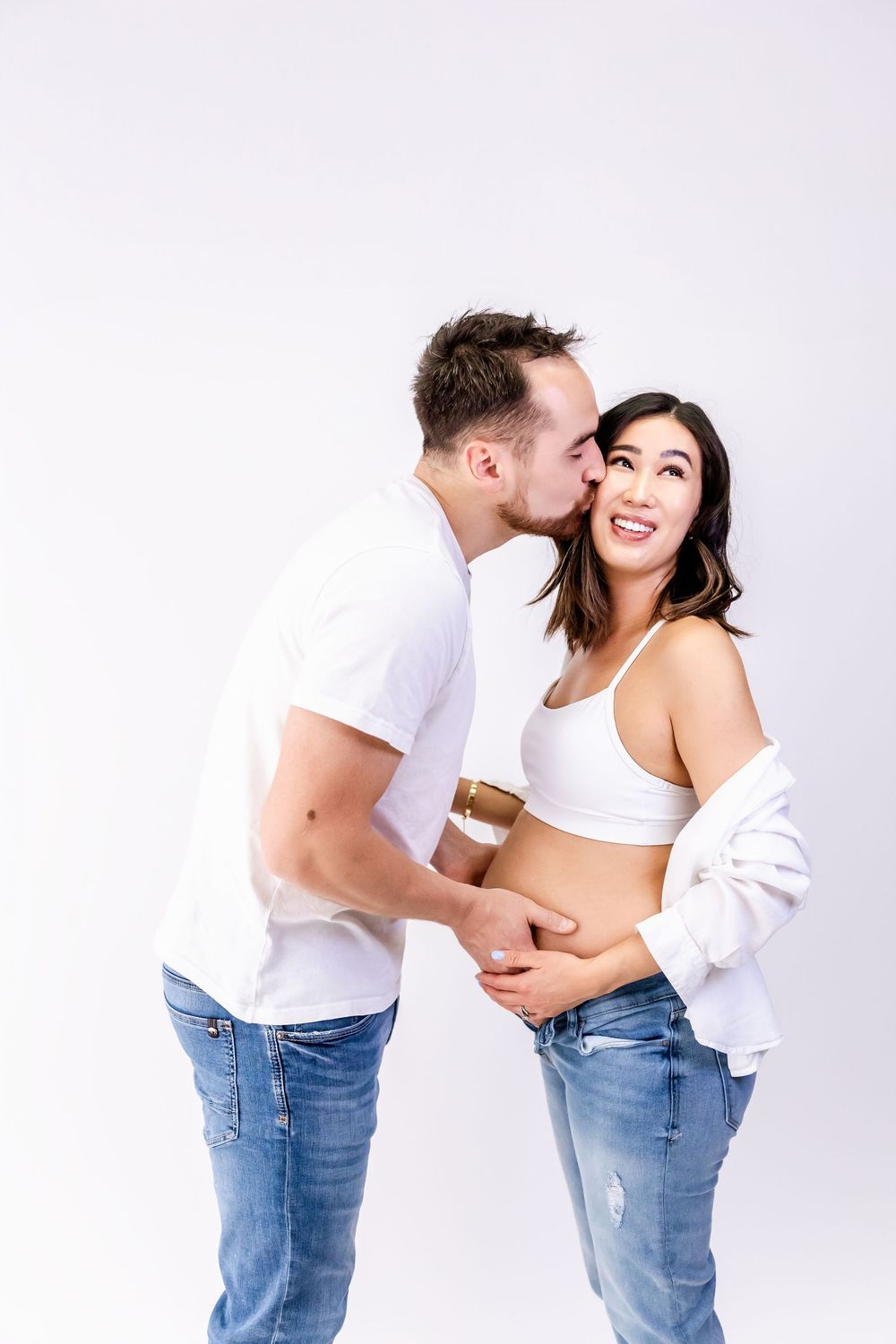 Joyful couple in matching white tops and blue jeans sharing an intimate moment against white background.