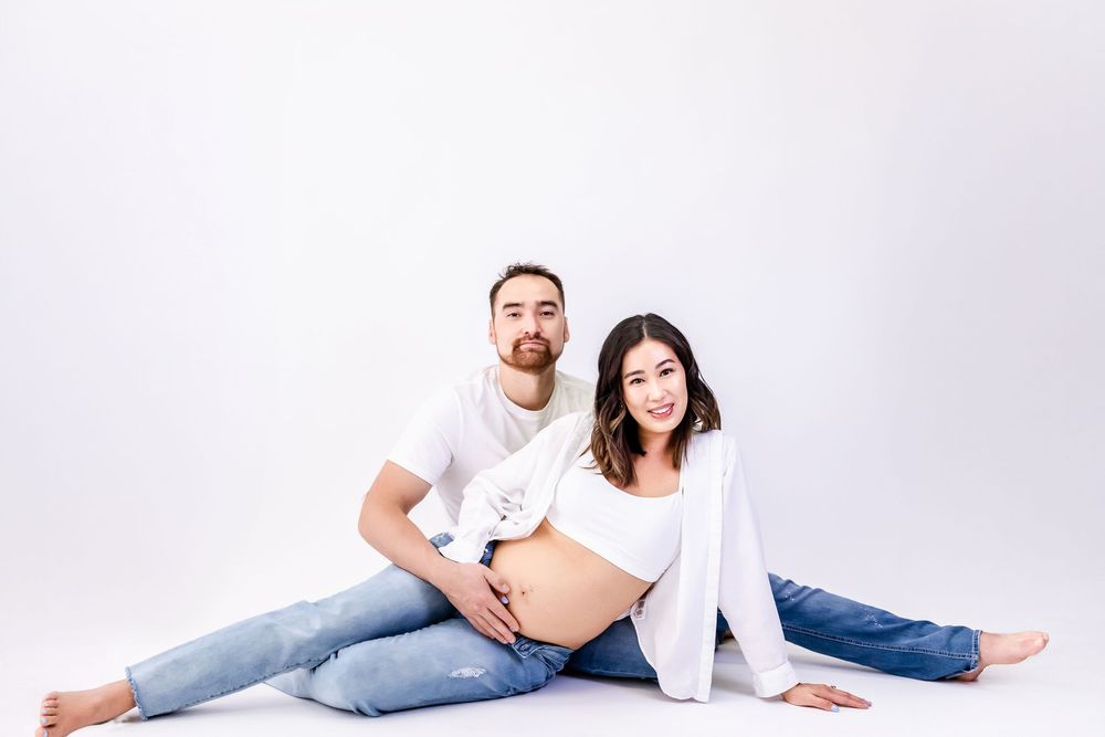 Expectant couple lounges together on white studio floor during intimate maternity photoshoot.